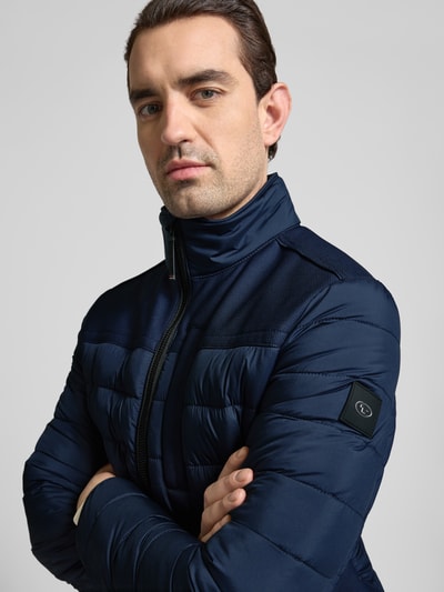 Mężczyzna nosi Regular Fit Steppjacke mit seitlichen Stretcheinsätzen von Tom Tailor, kolor granatowy, z logo, krój regular fit, stójka, zapinana na zamek, elastyczne mankiety i dół, wodoodporna, kieszenie na zamek, detal na ramionach, widok z przodu, zbliżenie na górną część ciała.