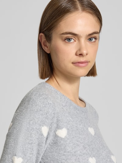 Modelka nosi sweter z dzianiny 'Strickpullover mit geripptem Saum' od REVIEW, elastyczna mieszanka materiałów, prosty krój, okrągły dekolt, prążkowany dół, długość tyłu w rozmiarze S: 47 cm, długość rękawów w rozmiarze S: 64 cm, jasnoszary z białymi sercami, miękka faktura. Zbliżenie na górną część ciała z przodu.