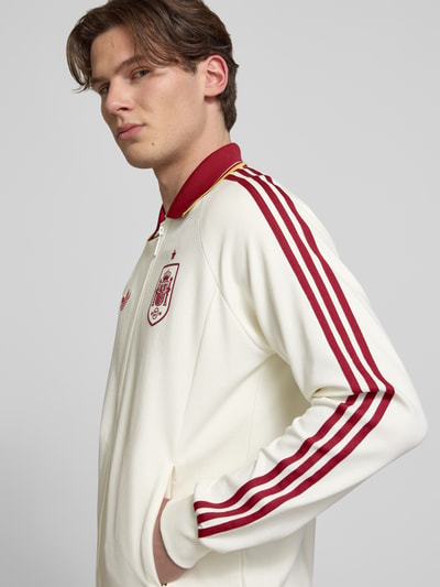 Männermodel trägt Regular Fit Trainingsjacke aus Baumwoll-Mix Modell 'BLACAS' von adidas Sportswear in Optik: Unifarben mit Struktur, Kontraststreifen, Logo-Stitching, Eingrifftaschen und Umlegekragen, Farbe Creme mit Rot, Ganzkörperansicht von vorne.