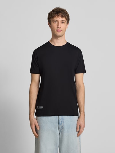 Męski model nosi T-Shirt mit Label-Patch od Alpha Industries, czarny kolor, bawełna, krój prosty, okrągły dekolt, widok całej sylwetki z przodu, długość tyłu w rozmiarze M: 66 cm.