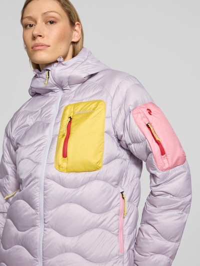 Frauenmodel trägt Jacke mit Entendaunen-Füllung und Kapuze von PEAK PERFORMANCE in Flieder mit gelben und rosa Taschen, prosty krój, kaptur z kordelstopperami, zamek błyskawiczny, wyhaftowane logo, brustasza zapinana na zamek błyskawiczny, kieszeń na rękawie, boczne kieszenie na zamek błyskawiczny, regulowane wykończenia, długość tyłu w rozmiarze S: 65 cm, szerokość ramion w rozmiarze S: 40 cm, szerokość w rozmiarze S: 51 cm, długość rękawów w rozmiarze S: 63 cm. Nahaufnahme Oberkörper