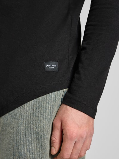 Mężczyzna nosi Longsleeve z prążkowanym dekoltem model 'ENOA' od JACK & JONES, bawełna, widoczne logo, zbliżenie na dolną część tułowia.