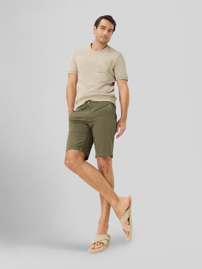 Männermodel trägt Regular Fit Chinoshorts aus Baumwoll-Mix Modell 'BOZEN' von Brax in Olivgrün, Kombination mit hellem Shirt, Ganzkörperansicht von vorne.