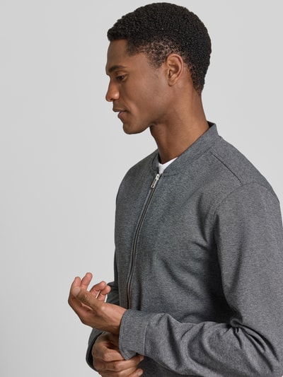 Męski model nosi Regular Fit Sweatjacke mit Stehkragen von Lindbergh, szary, gładka faktura, krój regular fit, zamek, kieszenie, elastyczny materiał, długość tyłu 69 cm, długość rękawa 66 cm, półzbliżenie, widok z boku.
