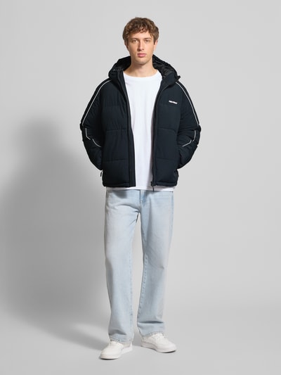 Männermodel trägt schwarze Steppjacke mit Kapuze von REVIEW, Regular Fit, Label-Print, Reißverschlusstaschen, Ganzkörperansicht von vorne.