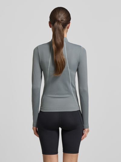 Frauenmodel trägt Longsleeve mit Stehkragen und Reißverschluss von Under Armour, Material-Mix mit Stretch-Anteil, Slim-Fit, Logo, Rückenlänge bei Größe S: 52 cm, gedeckte Farben, glatte Textur, Ganzkörperansicht Rückansicht.