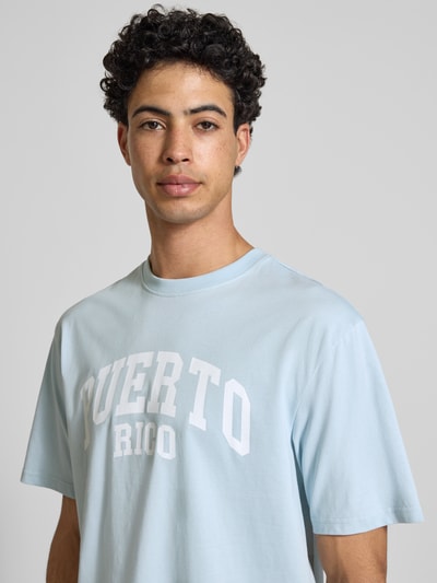 Männermodel trägt Relaxed Fit T-Shirt aus reiner Baumwolle Modell 'RICO' von Only & Sons, hellblau mit weißem Motiv-Print, Nahaufnahme Oberkörper.