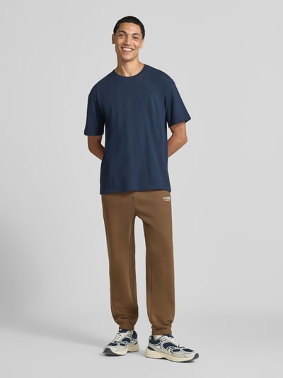 Männermodel trägt Straight Fit Sweatpants aus Baumwoll-Mix Modell 'Kane' von JACK & JONES in Braun mit Logo-Detail, Ganzkörperansicht von vorne. Herren Sweatpants im Straight Fit mit elastischem Bund und Gesäßtasche, Innenbeinlänge 72 cm.