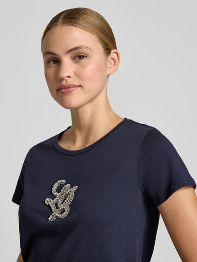 Frauenmodel trägt T-Shirt mit Rundhalsausschnitt und Strasssteinbesatz von LIU JO SPORT in Dunkelblau, Logo-Detail, Nahaufnahme Oberkörper.