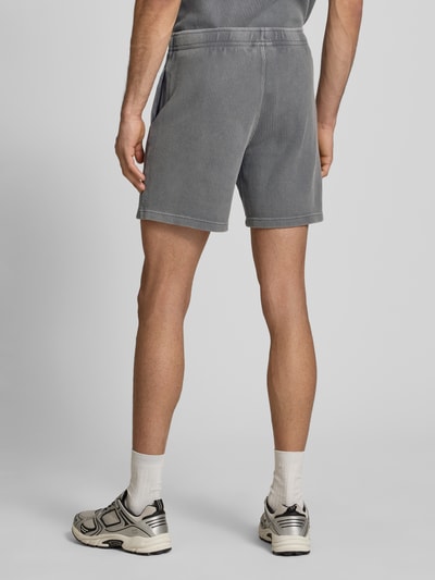 Męski model nosi Shorts mit Strukturmuster und elastischem Bund od CHAMPION, bawełniany miks, krój regular fit, szare, teksturowane, label-stitching, szerokość w rozmiarze M 40 cm, długość nogawki wewnętrznej w rozmiarze M 19 cm, widok całej sylwetki z tyłu.