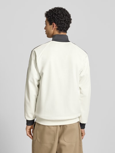 Männermodel trägt Regular Fit Trainingsjacke mit Vereins Logo Applikation Modell 'BLACAS' von adidas Originals, beige mit schwarzen Kontraststreifen, gerippte Abschlüsse, Stehkragen, unifarben, ganzkörper Ansicht Rückansicht.