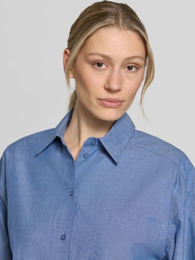 Kobiece model nosi Cropped Bluse aus reiner Bio-Baumwolle Modell 'KIRA-ASTHA' von Selected Femme, denimowa tkanina, kołnierzyk koszulowy, listwa guzikowa, długi rękaw, brustasza, czysta bawełna ekologiczna, efekt skróconego kroju, z przodu, zbliżenie na górną część ciała.
