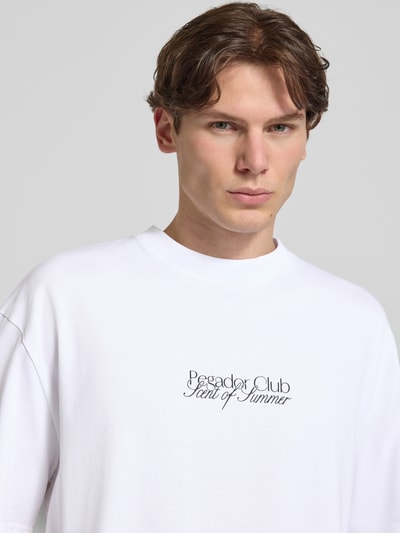 Męski model nosi Oversized T-Shirt mit Label-Print Modell 'Ramon' von PEGADOR z bawełny, biały, okrągły dekolt, nadruk z logo, zbliżenie na górną część ciała.