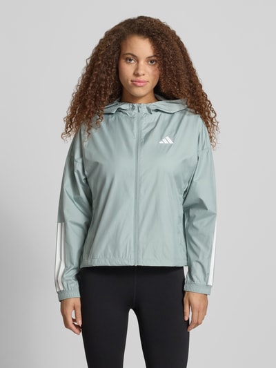 Modelka kobieca nosi Windbreaker mit Kapuze und Logo od adidas Sportswear, kolor pastelowy zielony, logo i zamki białe, krój relaxed fit, kaptur, boczne kieszenie, długość tyłu w rozmiarze S: 58 cm, szerokość ramion w rozmiarze S: 66 cm, szerokość klatki piersiowej w rozmiarze S: 53 cm, długość rękawa w rozmiarze S: 50 cm, widok całej sylwetki z przodu.