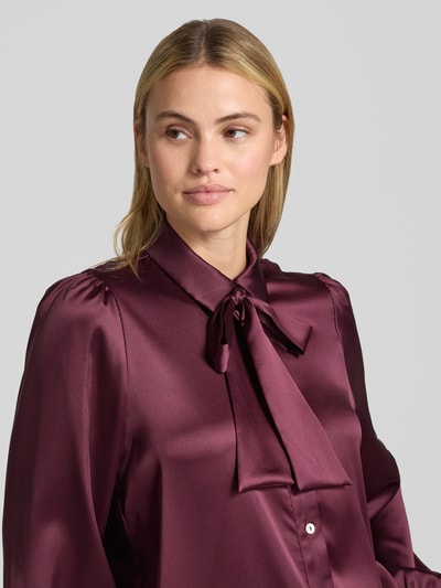 Vrouwenmodel draagt Regular Fit Hemdbluse aus Satin van Christian Berg Woman Selection in bordeauxrood met sierstrik, satijnen textuur, blousekraag, lange mouwen, detailopname bovenlichaam.