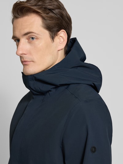 Mężczyzna nosi Parka z patkami przy mankietach model 'GEORGE' od Didriksons, granatowy, kaptur, krój regular fit, detaliczny widok na górną część tułowia, kontekst: długość tyłu w rozmiarze M: 94 cm, szerokość ramion w rozmiarze M: 45 cm, szerokość klatki piersiowej w rozmiarze M: 55 cm, długość rękawów w rozmiarze M: 67 cm.