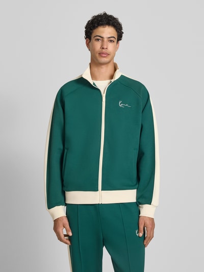 Männermodel trägt Regular Fit Sweatjacke mit Stehkragen von KARL KANI in Grün mit Kontrastdetails, Logo-Stitching, Reißverschluss, Eingrifftaschen, Elastischer-Material-Mix mit Baumwoll-Anteil, Ganzkörperansicht von vorne.