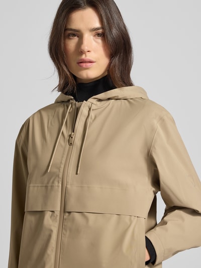 Frauenmodel trägt Regular Fit Jacke mit Kapuze Modell 'Suva' von Rains in Beige, Zweiwege-Reißverschluss, glatte Textur, Nahaufnahme Oberkörper. Rückenlänge 59 cm, Schulterbreite 49 cm, Brustweite 54 cm, Ärmellänge 60 cm.