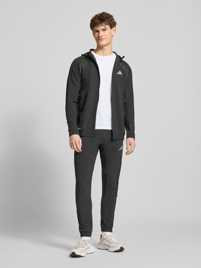 Männermodel trägt Regular Fit Trackpants mit elastischem Logo-Bund von adidas Training, unifarben, seitliche Reißverschlusstaschen, Ganzkörperansicht von vorne.
