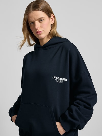 Kobiece model prezentuje bluza z kapturem z mieszanki bawełny, krój oversized fit, wyhaftowane logo, kieszeń kangurka, model 'No Hustle' od Karo Kauer, ciemnogranatowy, gładki materiał, z przodu, zbliżenie na górną część ciała.