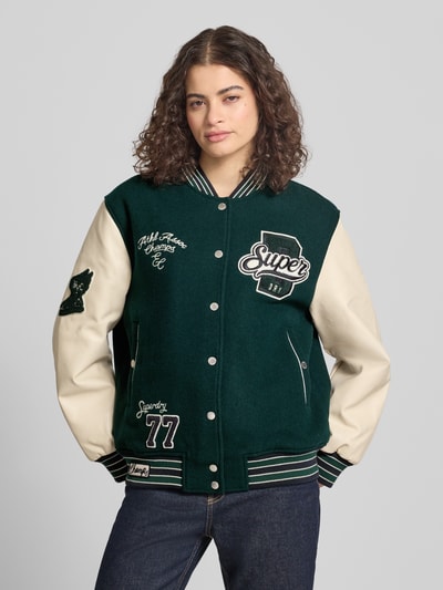 Frauenmodel trägt College Jacke mit Motiv- und Logo-Stitchings von Superdry in Dunkelgrün mit Woll-Mix, Lockerer-Schnitt, Stehkragen, Druckknopfverschluss, elastische Abschlüsse, Vorderansicht, Nahaufnahme Oberkörper. Rückenlänge 62 cm, Schulterbreite 49 cm, Brustweite 60 cm, Ärmellänge 60 cm, Größe S.