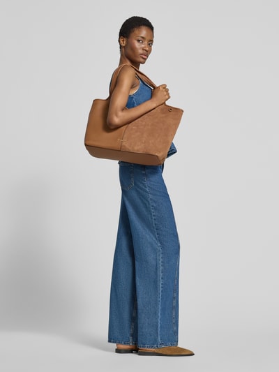 Frauenmodel trägt Tote Bag mit Logo-Applikation Modell 'Henly' von MICHAEL Michael Kors in Braun, große Rechteckform, glatte Textur, Hauptfach mit Magnetverschluss, Schulterriemen, Innenfach mit Reißverschluss, Seitenansicht, Ganzkörperansicht Rückansicht.