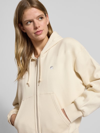 Modelka nosi bluzę rozpinaną z kieszenią kangurka Carhartt Work In Progress z bawełny, krój relaxed fit, kaptur, wyhaftowane logo, zamek błyskawiczny, kolor kremowy, widok z przodu, zbliżenie na górną część ciała.