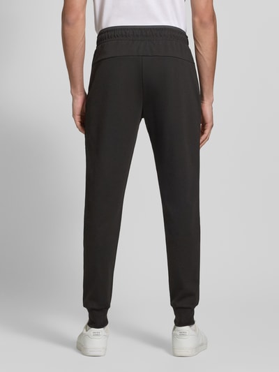 Männermodel trägt spodnie dresowe z elastycznej mieszanki materiałów z domieszką bawełny, model 'GORDON' od Jack & Jones, czarne, wąski krój, elastyczny pas z tunelem, elastyczne zakończenia nogawek, kieszenie zapinane na zamek błyskawiczny, długość wewnętrzna nogawki w rozmiarze M: 73 cm, widok całej sylwetki z tyłu