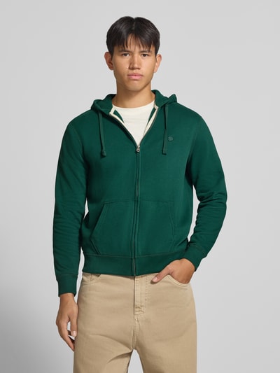 Männermodel trägt Regular Fit Sweatjacke aus Baumwoll-Mix mit Label-Detail von Superdry, Kängurutaschen, Kapuze, grün, Ganzkörperansicht von vorne.