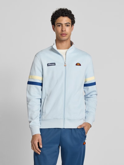 Männermodel trägt Sweatjacke aus Baumwoll-Mix Modell 'ROMA' von Ellesse in Hellblau mit Label-Details, Stehkragen, Eingrifftaschen, Gerade Passform, elastische Abschlüsse, durchgehender Reißverschluss; Rückenlänge 70 cm, Ärmellänge 68 cm, Ganzkörperansicht von vorne.