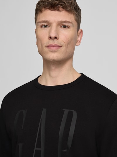 Männermodel trägt Sweatshirt mit Rundhalsausschnitt und Logo von GAP aus schwarzem Baumwoll-Mix, Regular-Fit, Nahaufnahme Oberkörper. Rückenlänge 69 cm, Ärmellänge 66 cm bei Größe M.