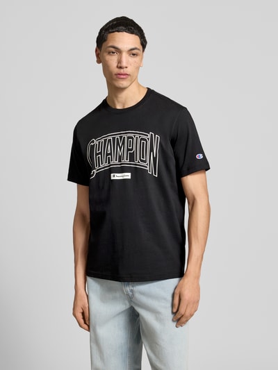 Männermodel trägt T-Shirt mit Logo und Rundhalsausschnitt von CHAMPION, schwarze Baumwolle, Regular Fit, Ganzkörperansicht von vorne.