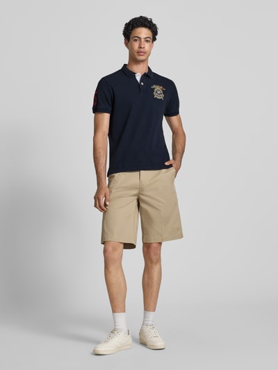 Männermodel trägt Regular Fit Poloshirt mit Label-Patches von Superdry in Dunkelblau, reine Baumwolle, kurze Knopfleiste, Ganzkörperansicht von vorne.