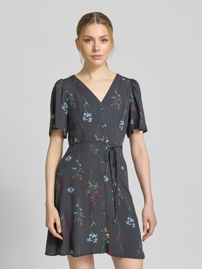Frauenmodel trägt Leicht ausgestelltes Minikleid aus reiner Viskose Modell 'ALBA' von Vero Moda in Dunkelgrau mit buntem Blumenmuster, V-Ausschnitt, kurzer weiter Ärmel, Knopfleiste, Taillengürtel, unifarbene Optik; Ganzkörperansicht von vorne.
