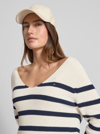 Frauenmodel trägt Regular Fit Strickpullover aus reiner Baumwolle von Tommy Hilfiger, Optik: Allover-Muster, gestreift, V-Ausschnitt, Langarm, Raglanärmel, Rippstrick, Logo-Stitching, Farbe creme mit navyblauen Streifen, Nahaufnahme Oberkörper.