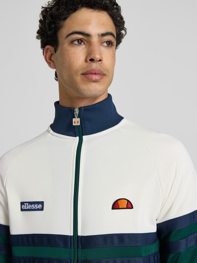 Männermodel trägt Sweatjacke aus Baumwoll-Mix Modell 'RIMINI' von Ellesse in Creme und Blau mit Label-Details, Nahaufnahme Oberkörper. Kontext: Herren Sweatjacke, Gerade Passform, Stehkragen, durchgehender Reißverschluss, elastische Abschlüsse, Rückenlänge bei Größe M 71 cm, Ärmellänge bei Größe M 43 cm.