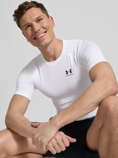 Mannelijk model draagt Slim Fit T-Shirt met Logo-Print van Under Armour, wit elastisch materiaal, slim fit, ronde hals, borstprint, close-up bovenlichaam.