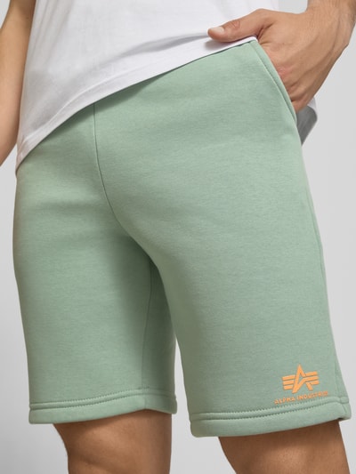 Männermodel trägt Shorts mit Eingrifftaschen und Logo-Print von Alpha Industries aus Baumwoll-Mix, Relaxed-Fit, mintgrün, Nahaufnahme Oberschenkel.