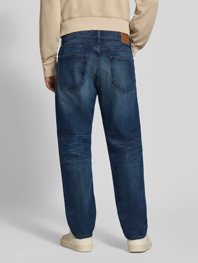 Männermodel trägt Tapered Fit Jeans aus Baumwoll-Mix von Tommy Jeans in Dunkelblau mit sichtbaren Nähten, Ganzkörperansicht Rückansicht. Herren Tapered Fit Jeans von Tommy Jeans.