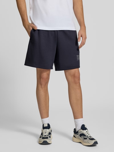 Männermodel trägt Sweatshorts aus Baumwoll-Mix von New Balance in Dunkelblau, gerader Schnitt, elastischer Bund, Label-Print, Nahaufnahme Unterkörper.