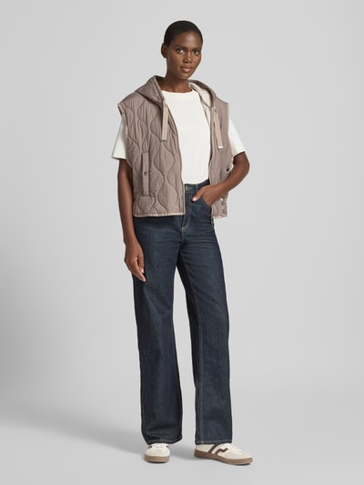 Frauenmodel trägt Steppweste mit Kapuze und Zweiwege-Reißverschluss von WHITE LABEL in Taupe, kombiniert mit Jeans, Regular Fit, seitliche Eingrifftaschen, matt strukturierte Oberfläche, Ganzkörperansicht von vorne.