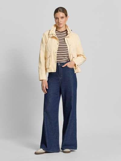Vrouwenmodel draagt Jacke mit Stehkragen und Pattentaschen van Fuchs Schmitt in regular fit, met tunnelkoord, knoopsluiting en klepzakken, beige kleur, gecombineerd met een gestreept shirt en wijde blauwe broek, geheel lichaamsaanzicht van voren.