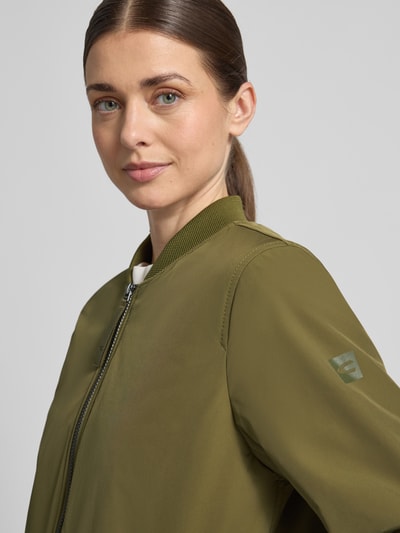 Frauenmodel trägt Jacke mit Zweiwege-Reißverschluss von camel active in Olivgrün, Stehkragen, elastische Ärmelabschlüsse, Nahaufnahme Oberkörper. Lockere Passform, extralanges Design, Details: Rückenlänge 92 cm, Schulterbreite 42 cm, Brustweite 55 cm, Ärmellänge 64 cm.