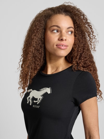 Modelka kobieca nosi Regular Fit T-Shirt mit Logo-Print Modell 'Loa' od Mustang, czarna bawełna, krótki rękaw, okrągły dekolt, zbliżenie na górną część ciała
