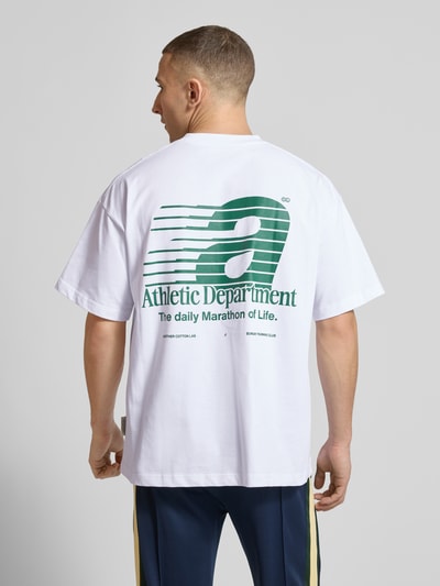 Męski model nosi t-shirt z nadrukiem z napisem model 'Athletic Department' od ANOTHER COTTON LAB, bawełna, prosty krój, okrągły dekolt, długość tyłu w rozmiarze M: 69 cm, widok całej sylwetki od tyłu.