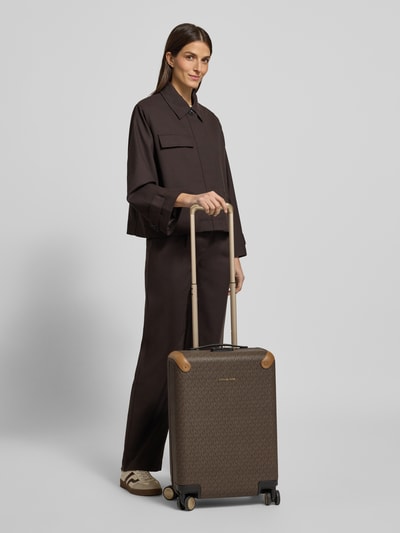 Frauenmodel trägt Trolley mit Label-Applikation von MICHAEL Michael Kors, zamek błyskawiczny dwustronny, teleskopowy uchwyt, szerokie 35 cm, długość 50 cm, głębokość 22 cm, aplikacja z metką, torba podróżna, uchwyty, brąz, prostokątne kształty, gładka tekstura, główna przegroda, kieszeń zapinana na zamek błyskawiczny, widok z przodu, całkowita sylwetka.