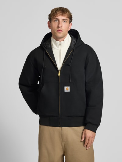 Männermodel trägt schwarze Jacke mit Label-Patch und Känguru-Tasche von Carhartt Work In Progress, Reißverschluss, Nahaufnahme Oberkörper.