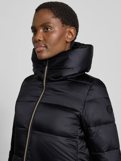 Frauenmodel trägt schwarzen Steppmantel mit Kapuze Modell 'LYSA' von Save The Duck, glänzende Oberfläche, Zweiwege-Reißverschluss, Label-Patch, Nahaufnahme Oberkörper.