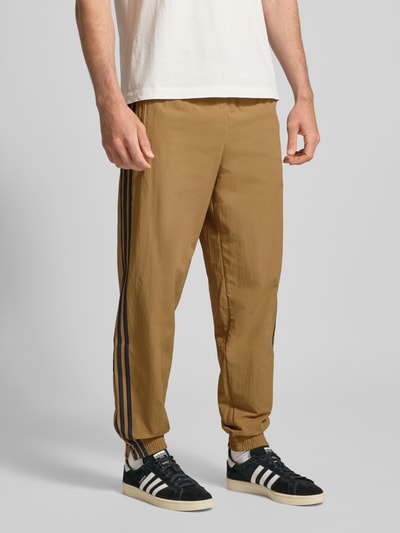 Mężczyzna nosi Sweatpants mit elastischem Bund od adidas Originals, luźny krój, elastyczny pas, beżowy kolor, widok całej sylwetki z przodu.