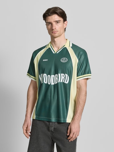 Männermodel trägt Poloshirt mit Label-Print Modell 'Taka' von WOODBIRD in Grün mit Polokragen, lockere Passform, Kurzärmelig, Ganzkörperansicht von vorne.